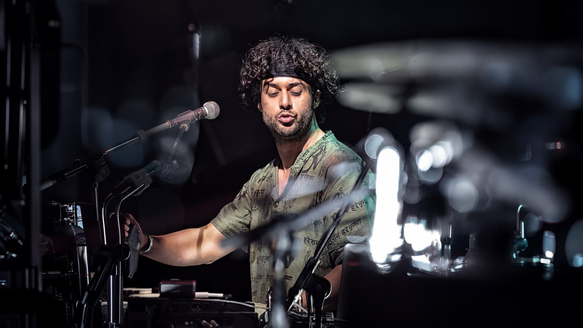 Osman Meyredi singing at the keys under stage light, black headband, dark stage